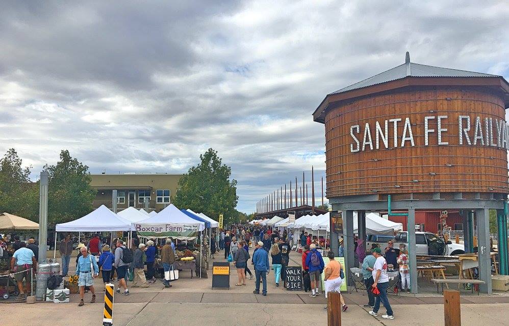 Busy outdoor market at the Santa Fe Railyard with tents, vendors, and visitors, next to the iconic wooden water tower bearing the 'SANTA FE RAILYARD' sign.