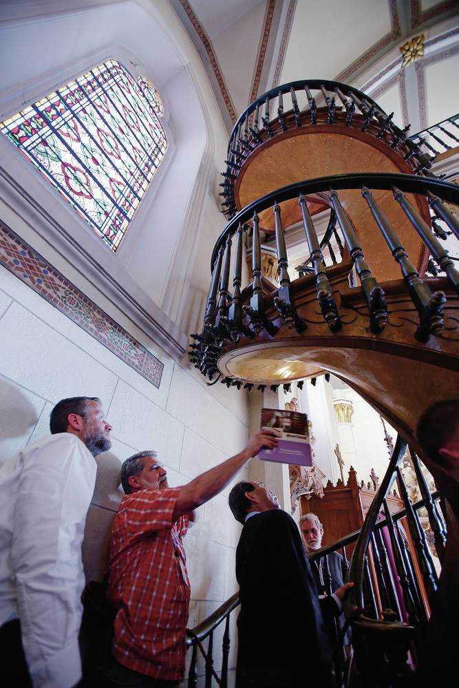 Photo of a group of people observing a historic spiral staircase in a church, with an architect leading the discussion about the design and restoration.